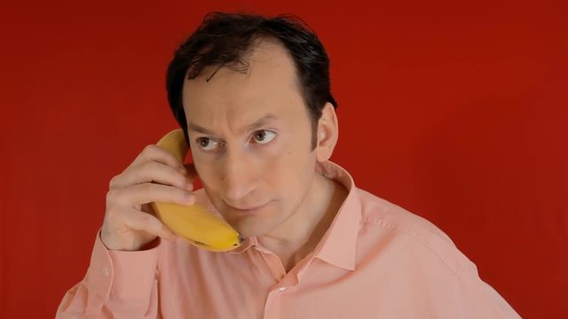 A funny ugly man talking on a banana phone, getting suspicious but being reassured. Red background.
