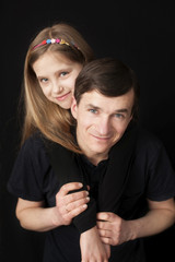 Daughter hugs her dad from behind on a black background. They both smile and look very happy. Vertical