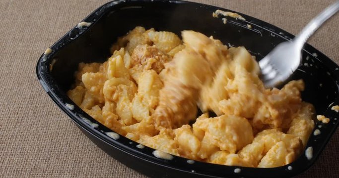 Close video of a spicy chicken nuggets with macaroni and cheese TV dinner in a black tray on a coarse brown tablecloth being eaten.