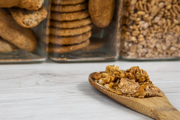 A wooden spoon full of granola on a white wood table, jars in the background full whit granola and biscuits