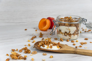 A  jar filled with granola and yogurt, wooden spoon whit granola and apricot on white wood table