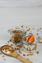 A  jar filled with granola and yogurt, wooden spoon whit granola and apricot on white wood table