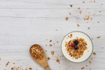 Bowl of yogurt with granola and fresh berries and wood spoon full whit granola on white wood table, background ,top view, copy space