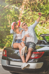 Young couple taking selfie while sitting at the back of their convertible car in nature. Road trip concept.