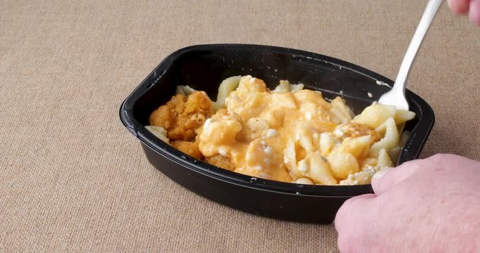 Video of a spicy chicken nuggets with macaroni and cheese TV dinner in a black tray being stirred after being microwaved on a coarse brown tablecloth.