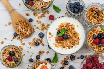 Jars full with granola, yogurt and fresh berries and wood spoon full whit granola on white wood table, top view, selective focus