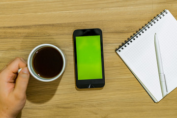 On the table is a smartphone and a notebook with a pen. The hand takes a cup of coffee from the table.