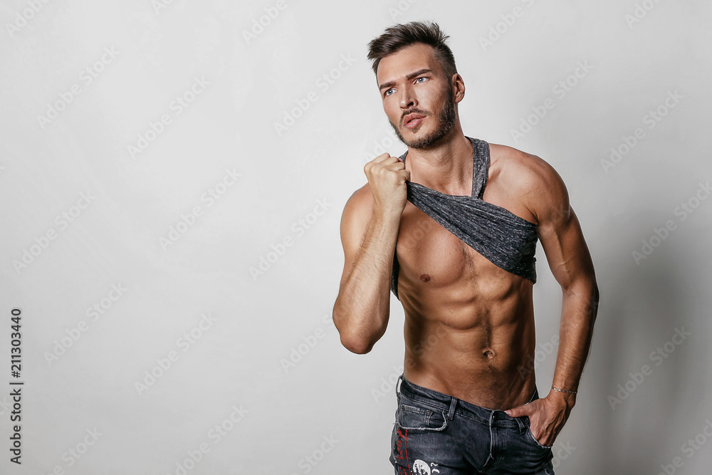 Wall mural muscular model sports young man in blue jeans raised his hand t-shirt and showing his press on a gre