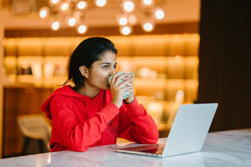 Young attractive Indian student enjoying a coffee whilst working with a laptop 