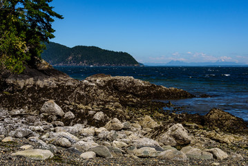 Fototapeta na wymiar Rocky shoreline on a beach in the San Juan Islands, the Salish Sea and other islands in the background, sunny day with a blue sky and while clouds 