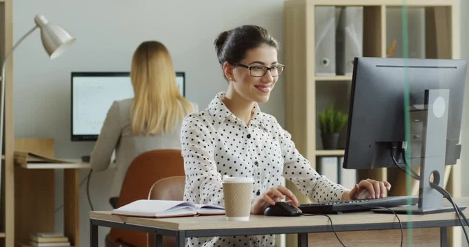 Young attractive female office worker using laptop computer while sitting in the office, then crossing out a task in the notebook ang going away. Indoors.