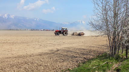 Tractor pulling heavy metal roller over dry field, with mountains, little bit snow on top, in background. Spring field preparation.