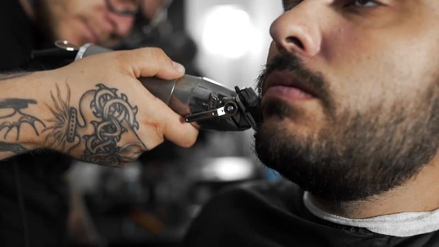 Tattoed barber shears the customer's beard by using trimmer at the barber shop, man's haircut and shaving at the hairdresser, shaving salon