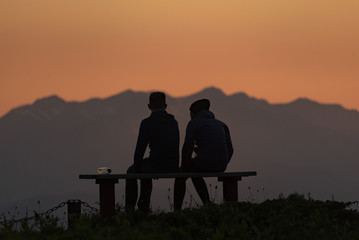 Fototapeta na wymiar Tourists rest on a bench in the mountains and admire the sunset over the mountain peaks.