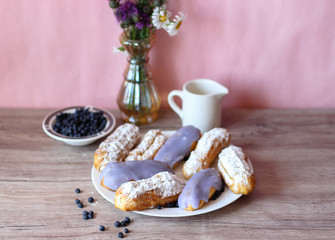 Berry glazed eclairs with blueberries on a wooden background with a decor consisting of a vase with wildflowers and a jug for milk and plate with blueberries