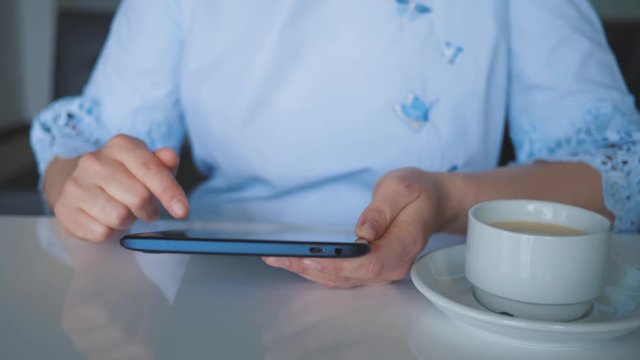 A young girl sitting at a table with a Cup of coffee, a digital tablet in her hands, leafing through the catalog of the online store on the touch screen of the tablet, cropped image.