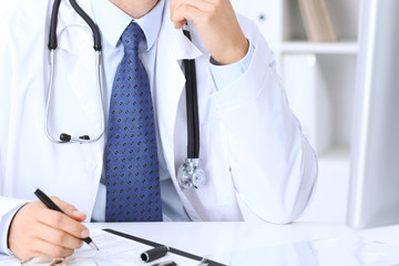 Unknown male doctor is sitting at the table and working in the hospital office. Closeup of stethoscope