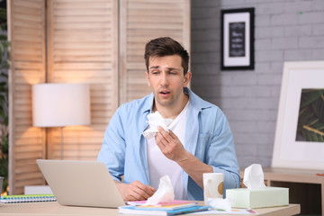 Sad exhausted man with tissue suffering from cold while working with laptop at table