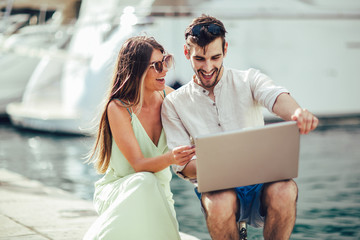 Beautiful romantic couple using laptop. Watching pictures on the laptop while traveling, by the harbor of a touristic sea resort with boats on background.