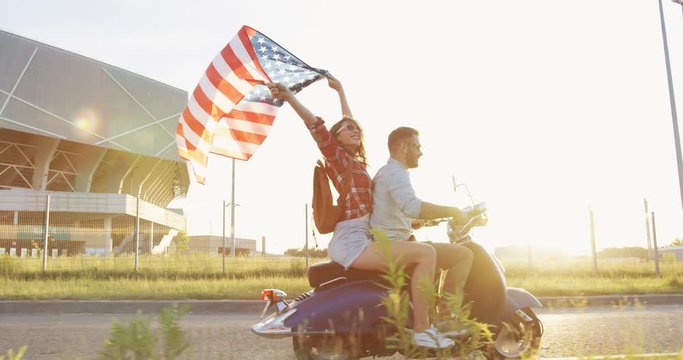 Side view on the young caucasian cheerful couple riding a motorbike and woman waving a big American flag near big stadium in the outskirts on a sunset. Outdoor.