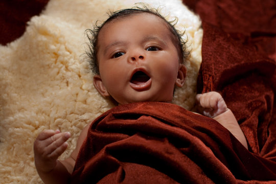 5 Wochen altes Baby mit dunkler Hautfarbe, schwarzen Haaren und Augen und ausdrucksstarkem Gesicht ist in ein braunes Tuch gehüllt, liegt auf einem Schafsfell und gähnt