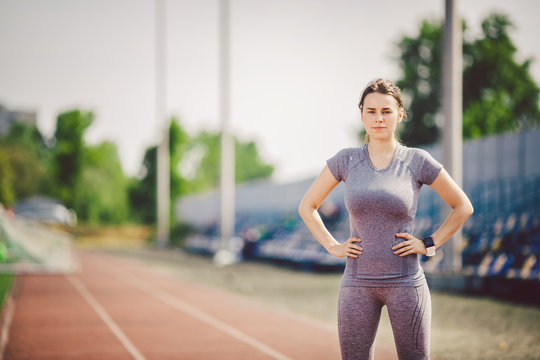 Portrait of a beautiful young caucasian woman with long hair in the tail and big breasts posing in gray sportswear standing training on a running stadium, a red rubber track in summer on a sunny day