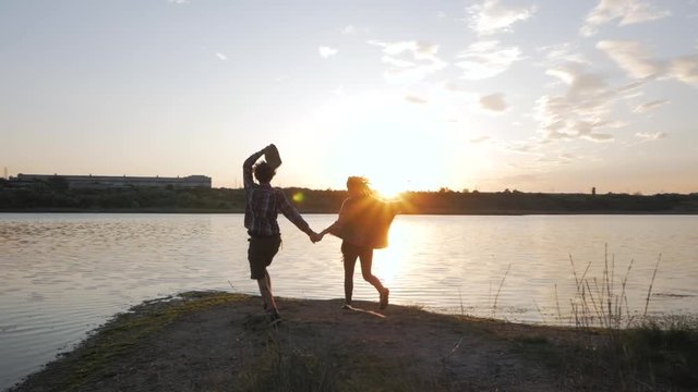 Young happy couple of lovers hiking near river in summer time against beautiful sunset