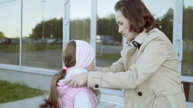 A woman zips up her daughter's sleeveless jacket. A woman tucks the sleeve of her daughter. A little girl with her mother in front of the station building.