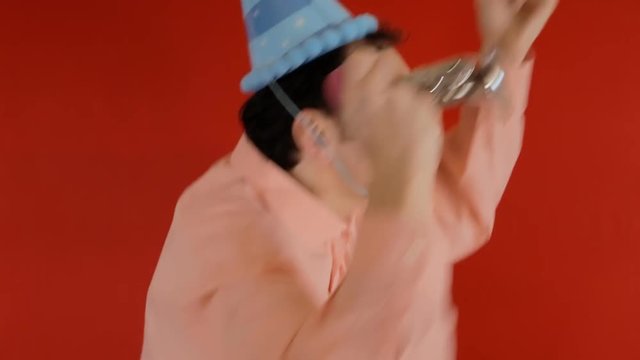 A funny ugly man feeling in good humor at a party, wearing a paper hat and squishing a horn, dancing, cheering. Red background.
