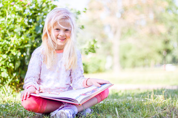 Fototapeta na wymiar blonde prodigy little girl is reading big book in garden at summer day and smiling at camera - education, knowledge, and people concept