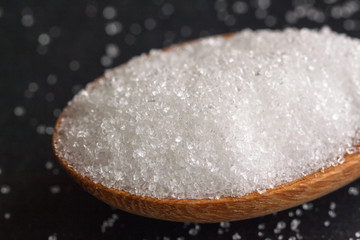 Natural purified sugar or bleach sugar on wood spoon on black granite table in close up view macro concept with copy space. Ingredient prepared for cooking or bakery. Unhealthy seasonings for diet.