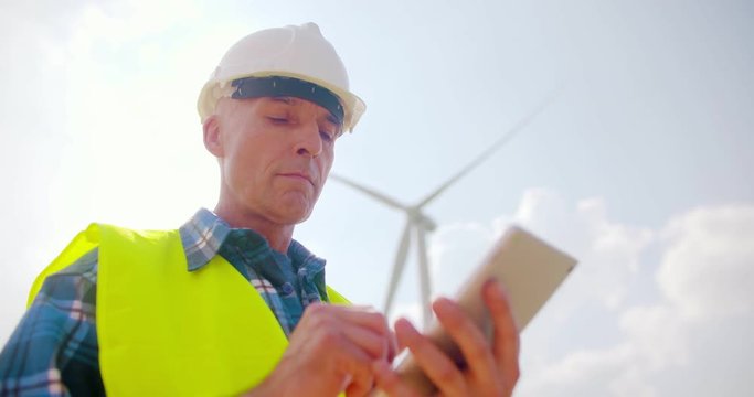 Smiling Engineer Using Digital Tablet At Windmill Farm Against Sky