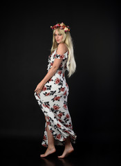 full length portrait of blonde girl wearing floral dress and a flower crown. standing pose against a black studio background.