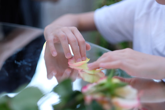 The hands of a child sitting at a table with a mirror surface and playing with rose petals.