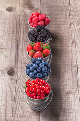 Berries fresh assortment in five tin cans stacked on rustic wooden table in studio. Strawberry, blueberry, blackberry, raspberry and red currant.