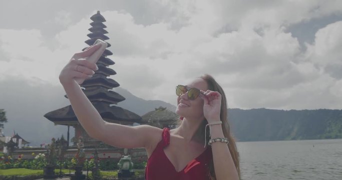 Young female tourist enjoys her holiday, doing selfie photo with her mobile phone at Ulun Danu Batur temple on the island of Bali, Indonesia - video in slow motion