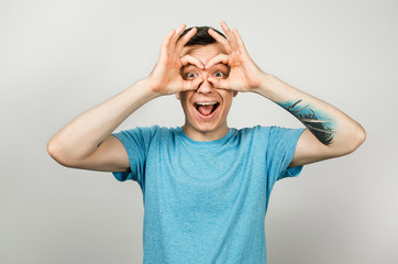 Young guy dressed in a blue t-shirt looks through binoculars from the palms of his hands, isolated on a light background.