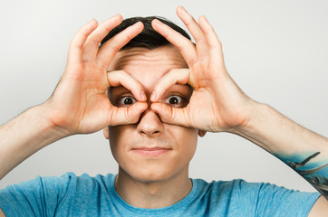 Young guy dressed in a blue t-shirt looks through binoculars from the palms of his hands, isolated on a light background. Close-up portrait