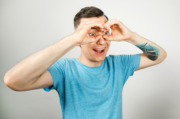 Young guy dressed in a blue t-shirt looks through binoculars from the palms of his hands, isolated on a light background.