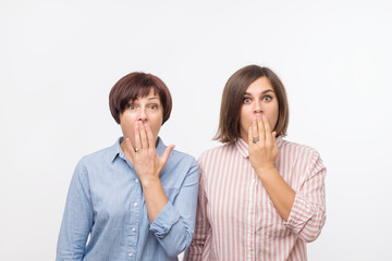 Portrait of a two surprised women mature mother and daughter standing over gray background and looking at camera. They are shocked of news and covering their mouths with plams
