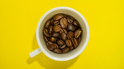 A small coffee cup filled with fried coffee beans. Yellow background. Top view