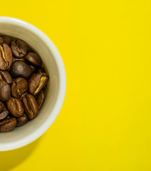 A small coffee cup filled with fried coffee beans. Yellow background. Top view