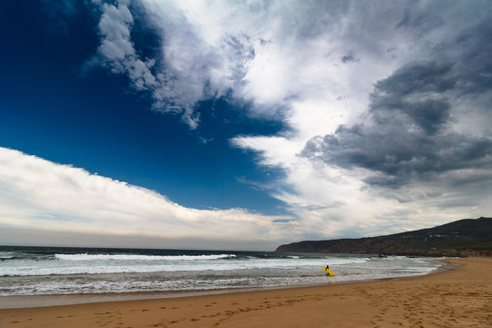 View of the Guincho beach near Atlantic coast. Surfer on the ocean coast in a wet suit with surfboard. Landscape of sunny day, blue sky and a mountain in distance. Cascais. Portugal.
