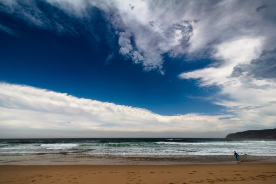 View of the Guincho beach near Atlantic coast. Surfer on the ocean coast in a wet suit with surfboard. Landscape of sunny day, blue sky and a mountain in distance. Cascais. Portugal.