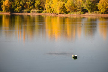 A calm autumn day, the fisherman's boat is in the middle of the lake. Yellow leaves of trees.