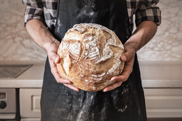 Hands of man holding big loaf of white bread. Male in black apron in home kitchen background with...