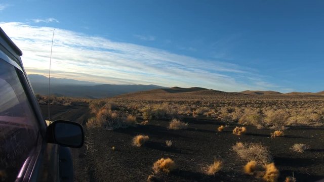 Stabilized camera movement over from outside a van at sunset golden hour. Black volcanic ground full of small rocks. Payun Liso volcano on background. Long shadows. Car window and rear view mirror.