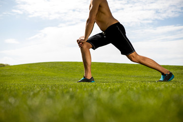 Athletic shirtless man is doing work out in fresh air. He is stretching with lunges and warming-up. Exercising in nature on warm day concept