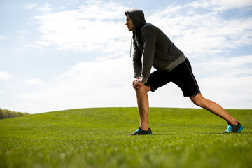 Concentrated man is doing lunges on green lawn during lovely day. He is stretching and warming-up on grass while looking ahead. Copy space in left side