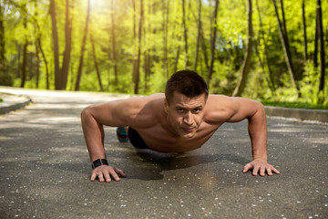 Obraz premium Athletic guy is concentrated on body workout. He is training pectorals with press-ups and wearing smartwatch. Male is exercising on road in green forest